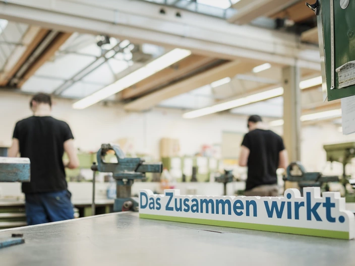 "Das Zusammen wirkt"-Schild in der Werkstatt mit zwei Azubis im Hintergrund